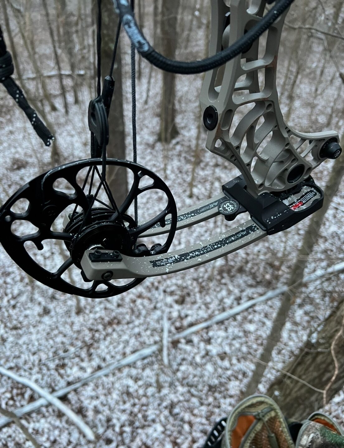 Close-up of a compound bow with snow on it, set against a snowy forest floor with bare trees in winter.