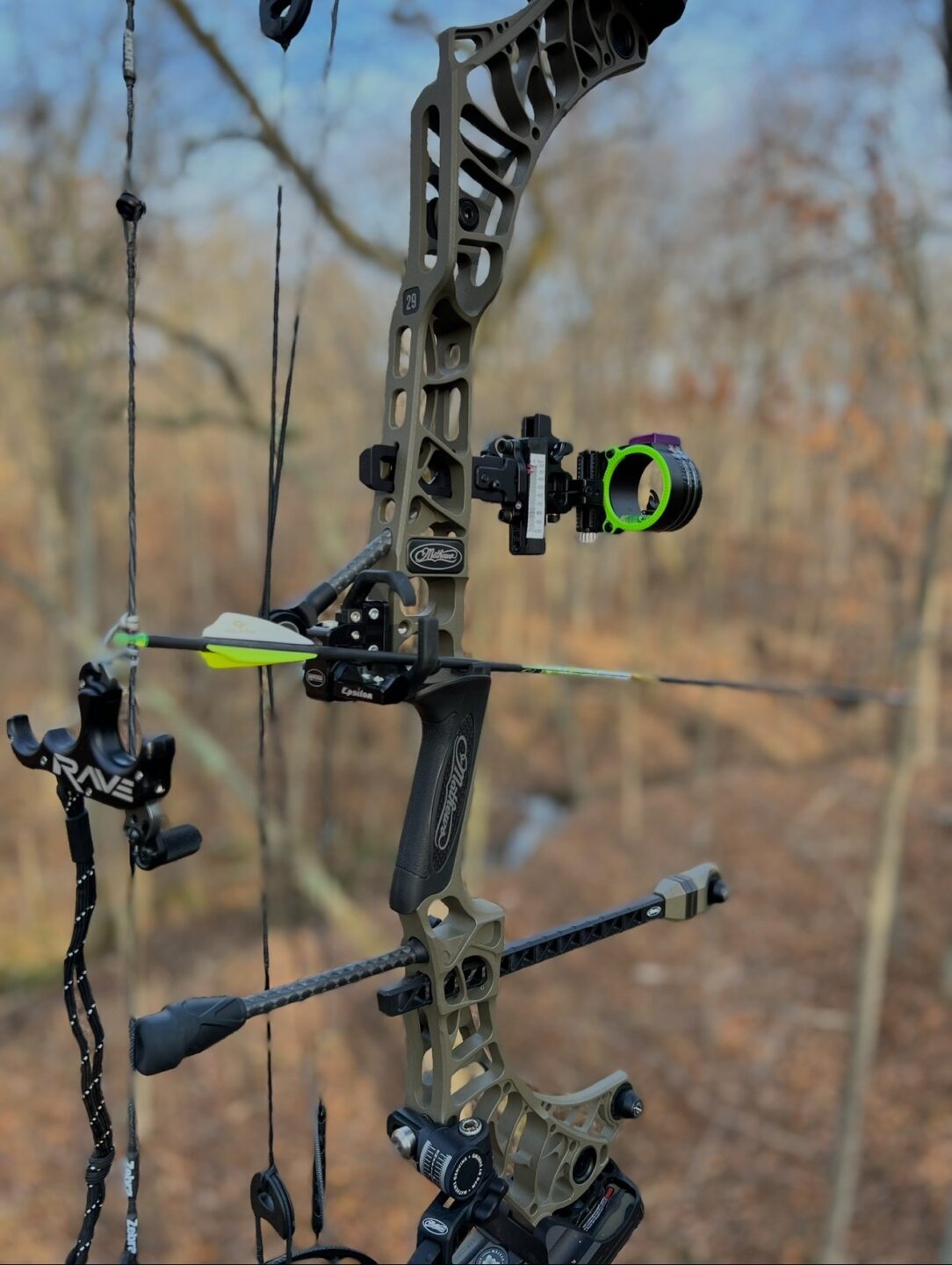 Close-up of a modern compound bow with an arrow nocked, equipped with sights and stabilizers, set against a blurred forest background.