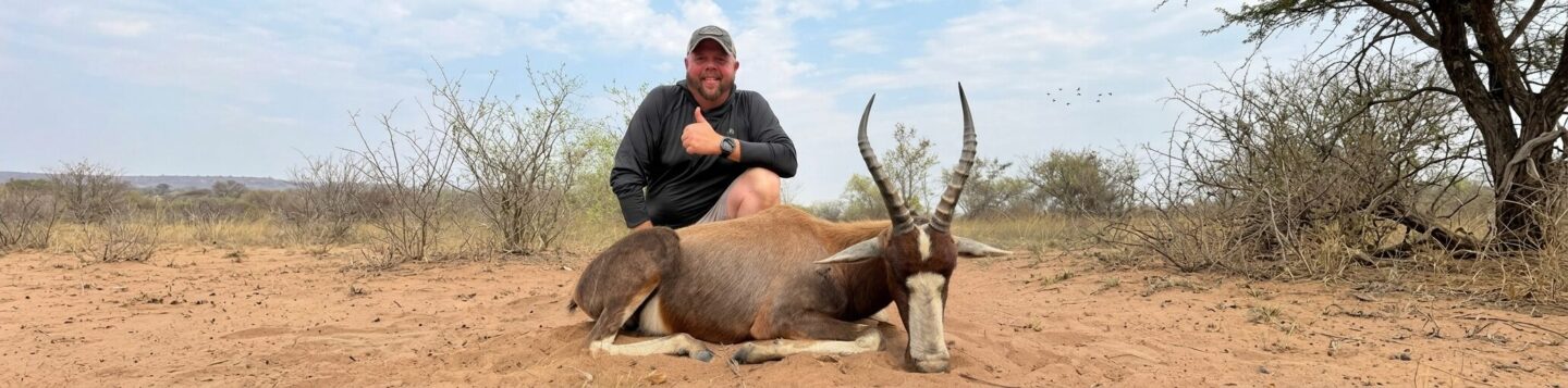 Man in outdoor clothing kneels on dry ground giving thumbs up next to a large antelope with long curved horns lying on the soil.